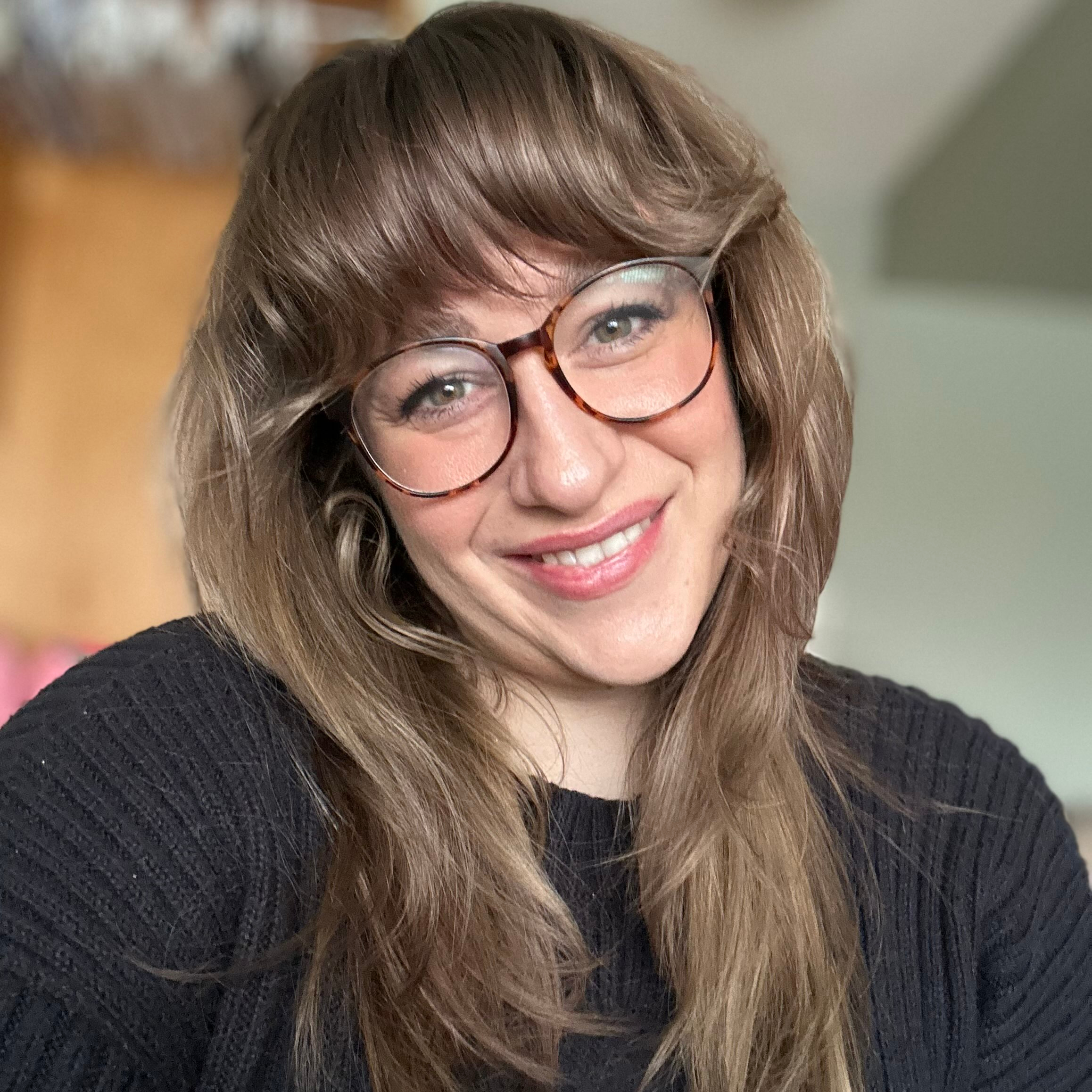 3llebelle smiles at the camera, head tilted to her right while wearing glasses and a black shirt.