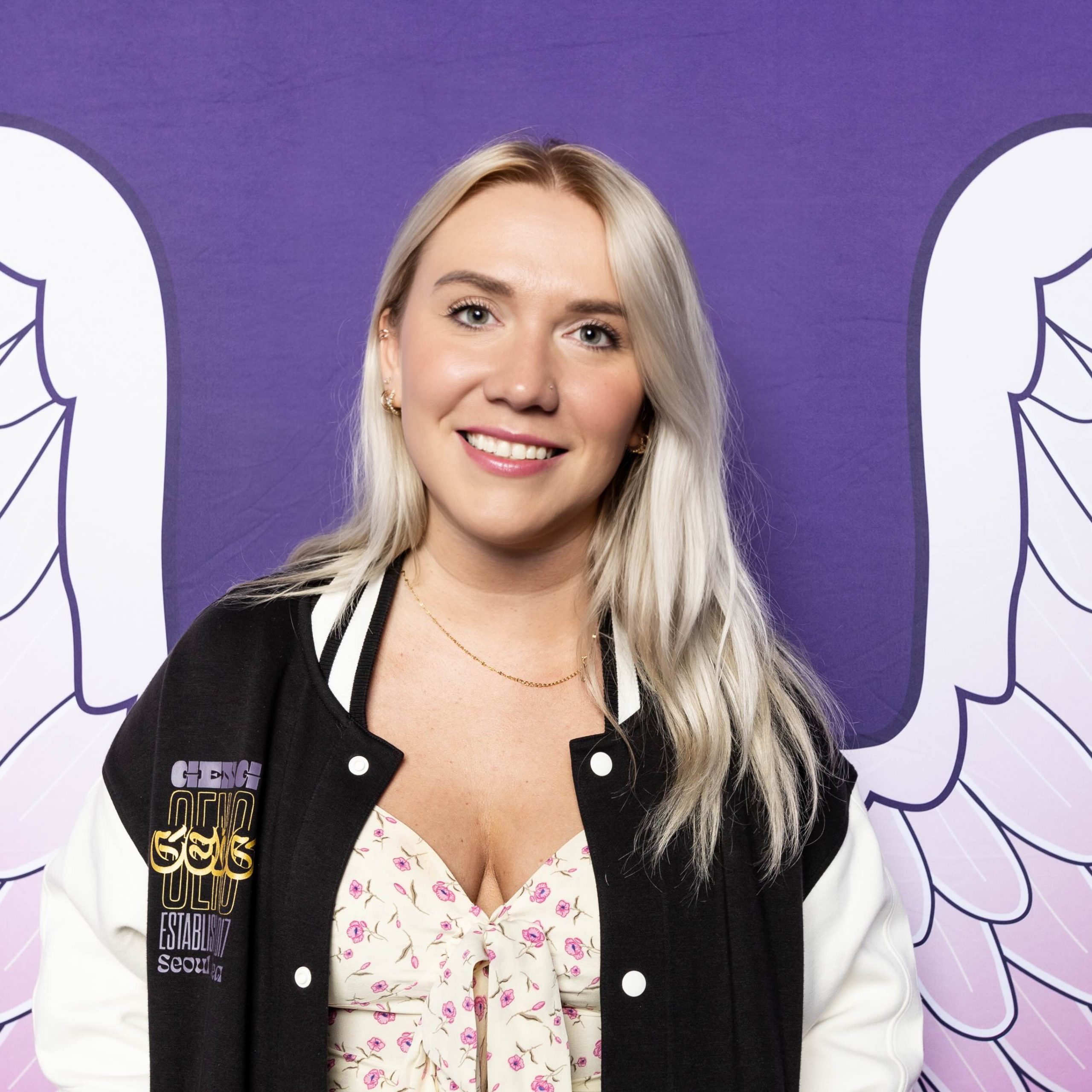 Widow wearing a black jacket and standing in front of a purple background with angel wings